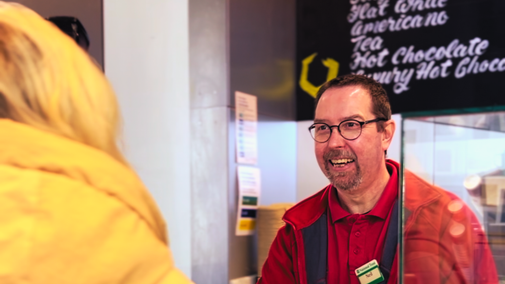 Visitor in a yellow puffer coat, standing at the till in the Giant's Causeway Visitor Centre Café, ordering with a team member named Neil. Signage in the background listing drinks and prices.